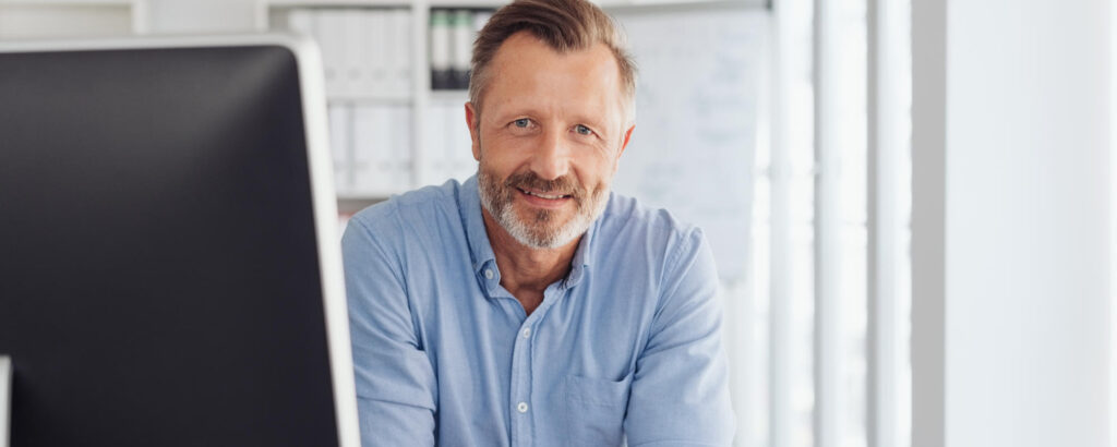 Banner. Attentive serious businessman seated at his desk peering around a desktop monitor staring at You